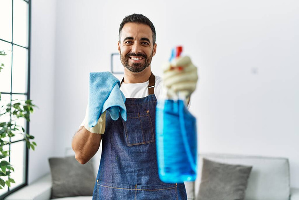Joven hombre hispano sonriendo confiado sosteniendo pulverizador en casa - Foto, Imagen