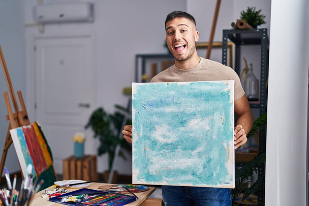 Νεαρός Ισπανός ζωγράφος κρατάει τον καμβά χαμογελώντας και γελώντας δυνατά γιατί αστειεύεται τρελά..  - Φωτογραφία, εικόνα
