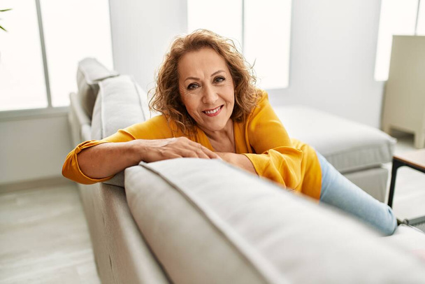 Donna caucasica di mezza età sorridente felice seduta sul divano di casa. - Foto, immagini