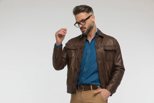 attractive man with one hand in pocket looking down and posing while wearing blue denim shirt with brown leather jacket in front of grey background - Photo, Image