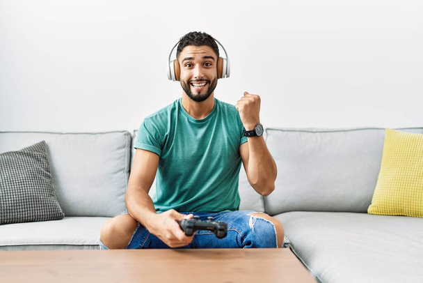 Handsome hispanic man wearing headphones playing video game holding controller screaming proud, celebrating victory and success very excited with raised arms  - Photo, Image