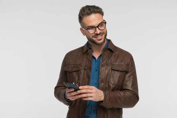glücklicher Mann mit Brille, der zur Seite schaut, während er in den sozialen Medien auf seinem Handy vor grauem Hintergrund im Studio scrollt - Foto, Bild