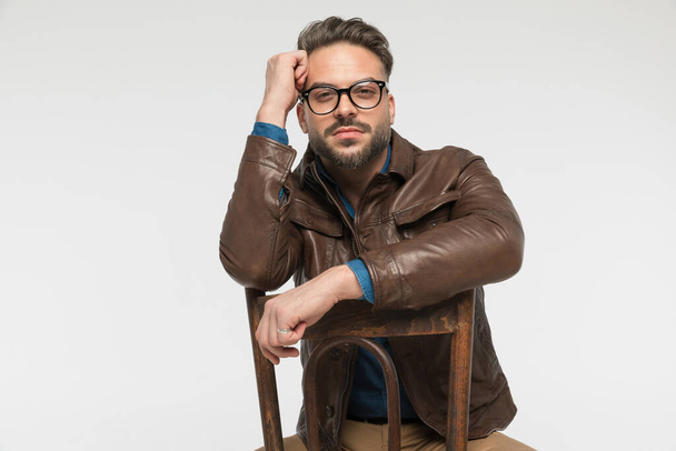 cool unshaved man in brown leather jacket with glasses holding arms and elbows on chair while sitting and posing on grey background - Fotografie, Obrázek