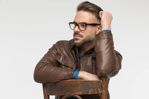 Gözlüklü, sandalyede dirsek tutan yakışıklı sakallı adam stüdyoda gri arka planda yer almak istiyor. - Fotoğraf, Görsel