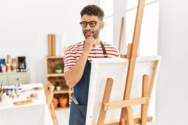 Hombre joven árabe en el estudio de arte con la mano en la barbilla pensando en la pregunta, la expresión pensativa. Cara sonriente y pensativa. concepto de duda.  - Foto, imagen