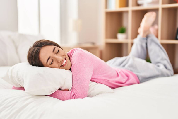 Mladý krásný hispánec žena objímání polštář ležící na posteli v ložnici - Fotografie, Obrázek