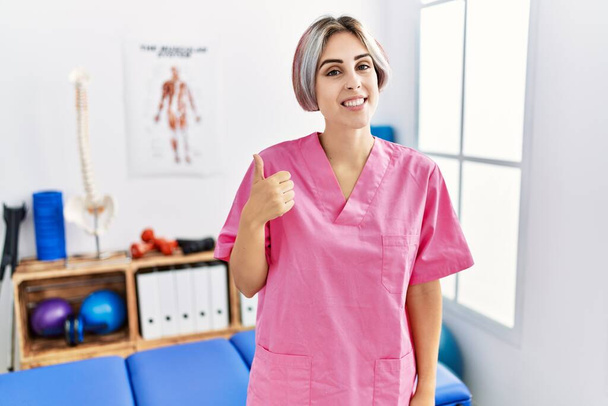 Jonge verpleegster werkt bij pijn herstel kliniek glimlachen gelukkig en positief, duim omhoog doen uitstekend en goedkeuring teken  - Foto, afbeelding