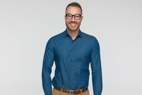young casual man is wearing a blue shirt and eyeglasses against gray studio background - Zdjęcie, obraz