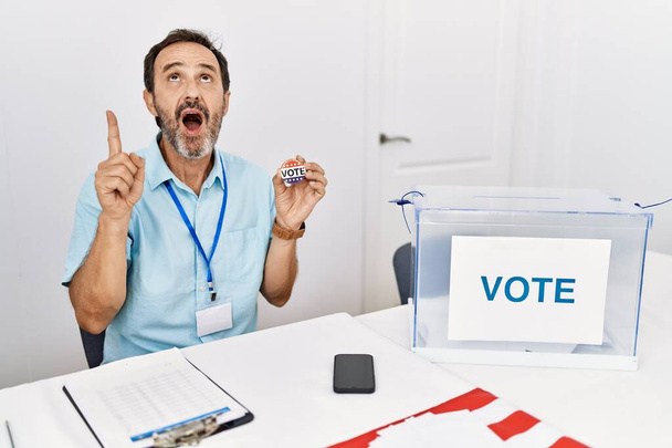 Hombre de mediana edad con barba sentada por papeleta sosteniendo voto insignia asombrado y sorprendido mirando hacia arriba y señalando con los dedos y los brazos levantados.  - Foto, Imagen