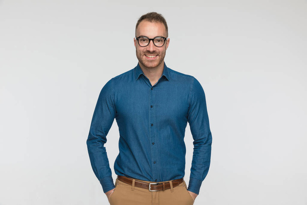 young casual man is posing with his arms in pocket and wearing a blue shirt and eyeglasses against gray studio background - Photo, Image