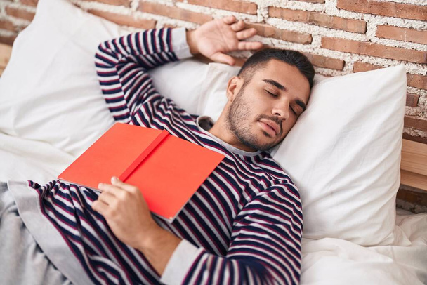 Young hispanic man holding book lying on bed sleeping at bedroom - Photo, Image