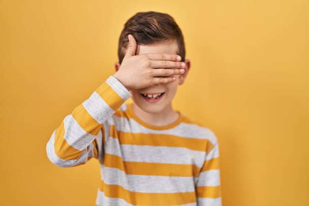 Young caucasian kid standing over yellow background smiling and laughing with hand on face covering eyes for surprise. blind concept.  - Zdjęcie, obraz
