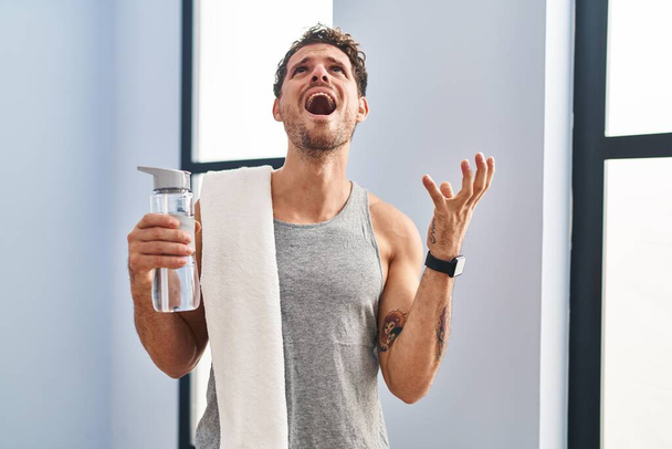 Young hispanic man wearing sportswear drinking water crazy and mad shouting and yelling with aggressive expression and arms raised. frustration concept.  - Photo, Image