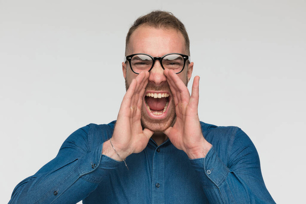 casual man is schreeuwen hardop met zijn handen rond zijn mond en het dragen van een blauw shirt en bril tegen grijze studio achtergrond - Foto, afbeelding