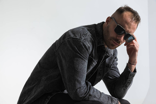 side view of cool young man with sunglasses holding elbow on knee and posing in front of grey background in studio - Foto, imagen