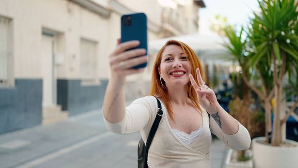 Junge rothaarige Frau macht Selfie mit dem Smartphone und macht Siegesgeste, mit den Fingern auf der Straße - Foto, Bild