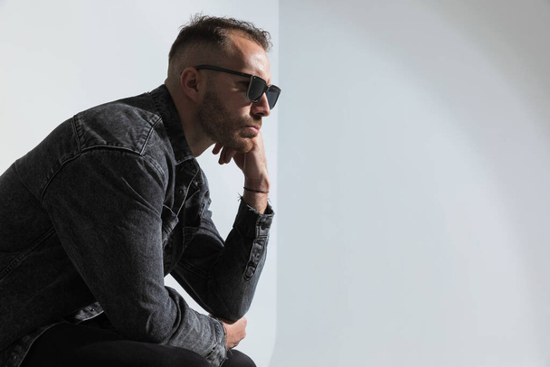 vista lateral del hombre casual reflexivo con gafas pensando y mirando al sidr, cogido de la mano a cara y sentado sobre fondo gris en el estudio - Foto, imagen