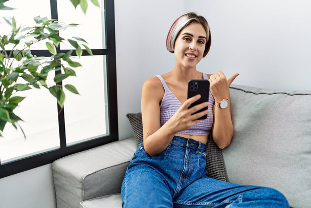 Mladá krásná žena pomocí smartphonu psaní zprávu sedí na pohovce ukazuje dozadu s rukou a palci nahoru, usmívá se sebevědomě  - Fotografie, Obrázek