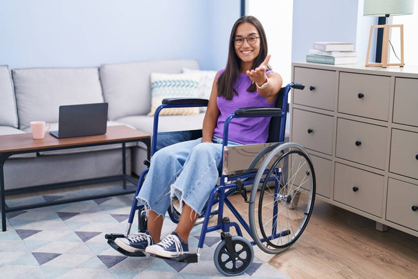 Joven mujer hispana sentada en silla de ruedas en casa sonriendo amigable ofreciendo apretón de manos como saludo y bienvenida. negocios exitosos.  - Foto, imagen