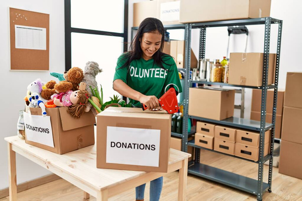 Mujer latina joven con uniforme voluntario embalaje caja de donaciones en el centro de caridad - Foto, imagen