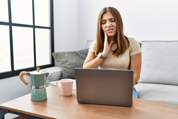 Młoda brunetka używająca laptopa w domu pijąca filiżankę kawy dotykającą ust z bolesnym wyrazem twarzy z powodu bólu zęba lub choroby zębów. dentysta  - Zdjęcie, obraz