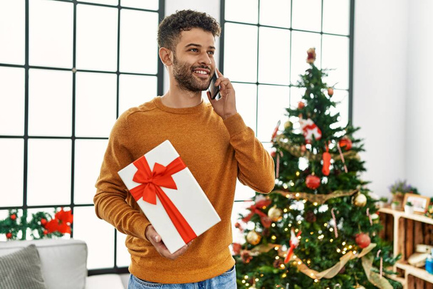 Junger Araber telefoniert mit dem Smartphone und hält Geschenk zu Hause am Weihnachtsbaum. - Foto, Bild
