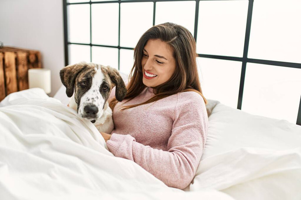 Junge Frau lächelt selbstbewusst und umarmt Hund, der auf Bett im Schlafzimmer sitzt - Foto, Bild