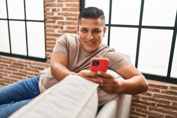 Young latin man using smartphone sitting on sofa at home - Foto, Bild
