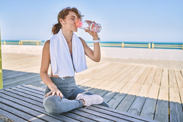 Молода біла жінка в спортивному одязі п'є воду на узбережжі
 - Фото, зображення