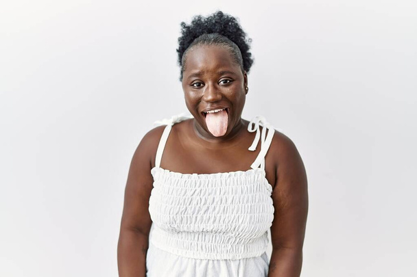 Junge Afrikanerin, die vor weißem, isoliertem Hintergrund steht, streckt die Zunge glücklich mit lustiger Miene heraus. Emotionales Konzept.  - Foto, Bild