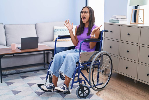 Νεαρή Ισπανίδα που κάθεται σε αναπηρικό καροτσάκι στο σπίτι τρελή και τρελή φωνάζοντας και φωνάζοντας με επιθετική έκφραση και χέρια σηκωμένα. έννοια απογοήτευσης.  - Φωτογραφία, εικόνα