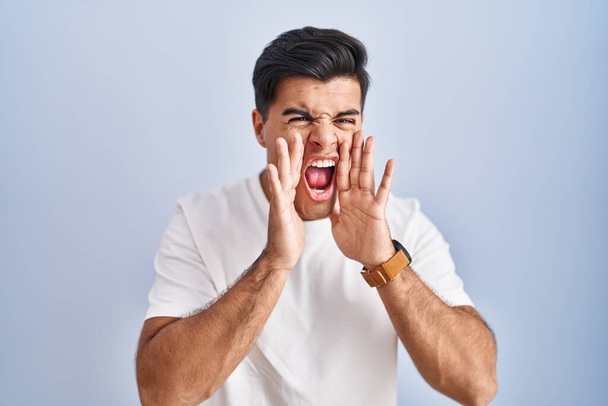 Spaanse man staat over blauwe achtergrond schreeuwen boos hardop met handen over de mond  - Foto, afbeelding