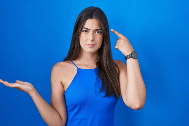 Испанская женщина, стоящая на синем фоне, смущена и раздражена открытой ладонью, показывающей пространство для копирования и указывающей пальцем на лоб. think about it.  - Фото, изображение