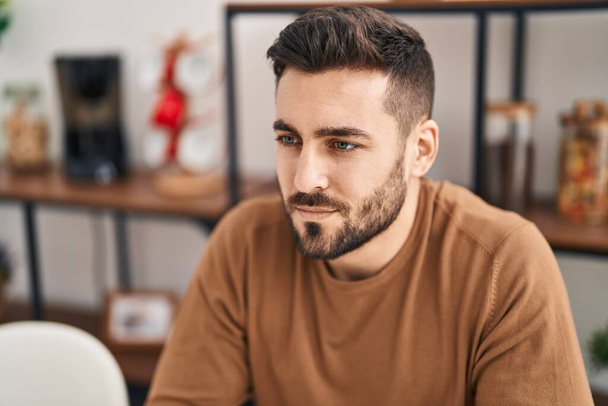 Young hispanic man sitting on table with relaxed expression at home - Foto, afbeelding