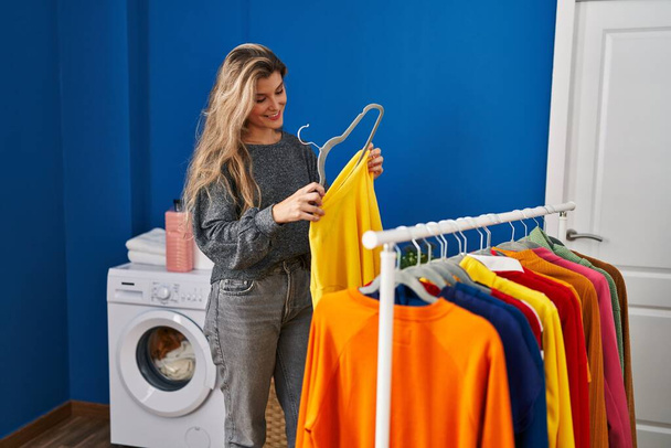 Young blonde woman smiling confident hanging clothes on rack at laundry room - Foto, afbeelding