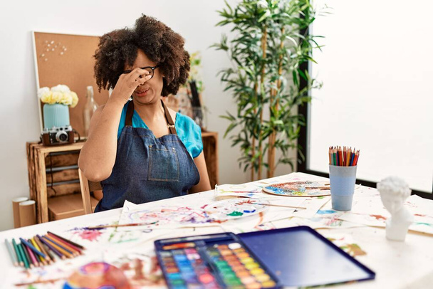 Hermosa mujer afroamericana con pintura afro en el estudio de arte oliendo algo apestoso y asqueroso, olor intolerable, conteniendo la respiración con los dedos en la nariz. mal olor  - Foto, Imagen