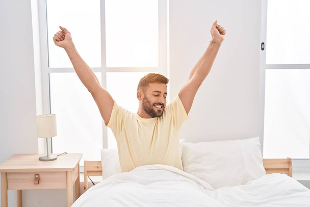 Young hispanic man waking up stretching arms at bedroom - Fotoğraf, Görsel