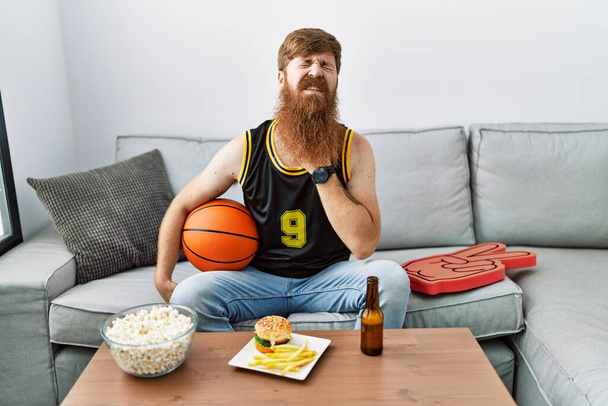 Homme caucasien à longue barbe tenant ballon de basket-ball acclamant jeu de télévision toucher le cou douloureux, mal de gorge pour la grippe, la motte et l'infection  - Photo, image