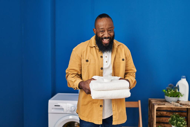 Afrikkalainen amerikkalainen mies pitää puhtaita pyyhkeitä pyykkihuoneessa hymyillen ja nauraen kovaa ääneen, koska hauska hullu vitsi.  - Valokuva, kuva