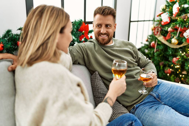 Νεαρό καυκάσιο ζευγάρι χαμογελά χαρούμενος και πίνοντας σαμπάνια κάθεται στον καναπέ από το χριστουγεννιάτικο δέντρο στο σπίτι. - Φωτογραφία, εικόνα