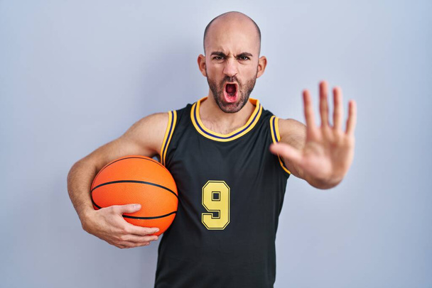 Jeune homme chauve avec barbe portant un uniforme de basket tenant le ballon faisant geste d'arrêt avec les mains paumes de la main, expression de colère et de frustration  - Photo, image