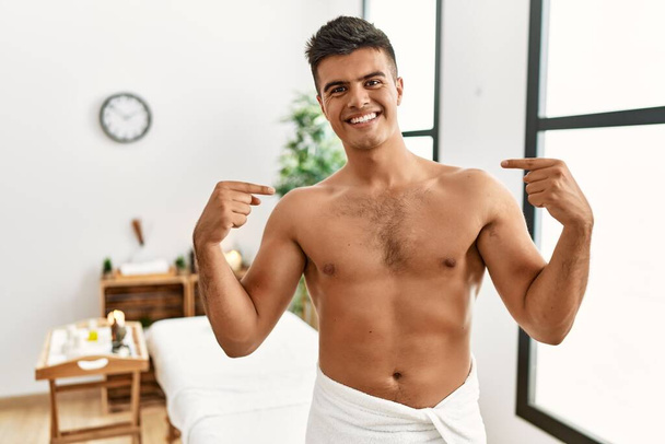 Jeune homme hispanique debout torse nu au centre de spa regardant confiant avec le sourire sur le visage, se montrant du doigt fier et heureux.  - Photo, image