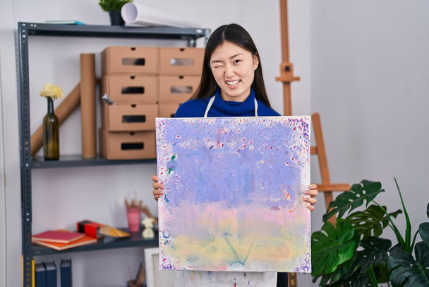 Chinesische junge Frau hält Leinwand im Kunststudio und blickt augenzwinkernd in die Kamera mit sexy Ausdruck, fröhlichem und glücklichem Gesicht.  - Foto, Bild