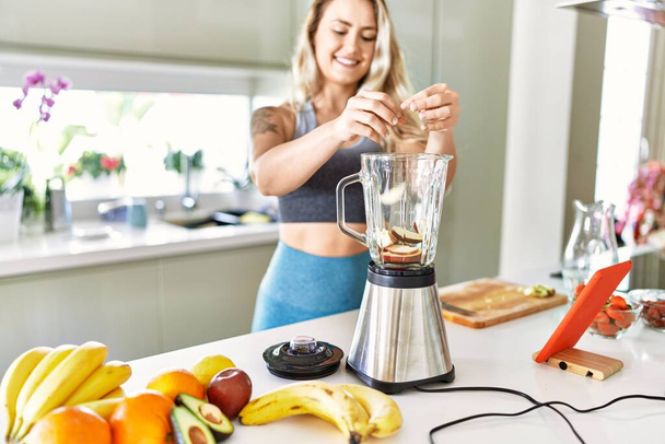Young woman looking online juice recipe pouring apple on blender at kitchen - Foto, imagen
