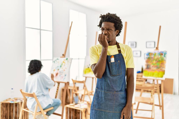 Afrikaanse jongeman die in de kunststudio staat en er gestrest en nerveus uitziet met handen op de mond bijtende nagels. angstprobleem.  - Foto, afbeelding