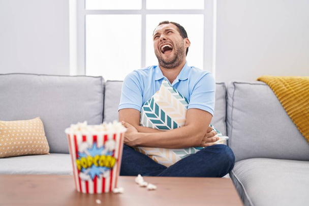 Spaanse man thuis tv aan het kijken zittend op de bank boos en boos schreeuwend gefrustreerd en woedend, schreeuwend van woede omhoog kijkend.  - Foto, afbeelding