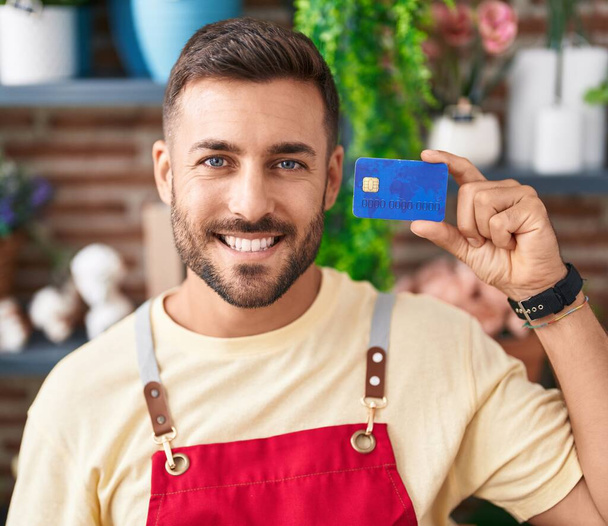 Çiçekçide çalışan yakışıklı İspanyol bir adam elinde kredi kartıyla mutlu ve pozitif görünüyor. Dişlerini gösteren bir gülümsemeyle ayakta duruyor.  - Fotoğraf, Görsel