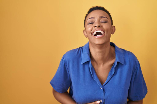 Afrikai amerikai nő áll sárga háttér mosolyog és nevetni keményen ki, mert vicces őrült vicc a kezét a test.  - Fotó, kép