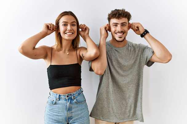 Mladý krásný pár stojí spolu nad izolovaným pozadím s úsměvem tahá za uši s prsty, legrační gesto. problém konkurzu  - Fotografie, Obrázek