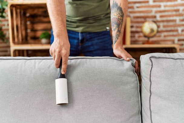 Young redhead man cleaning sofa using pet hair roller at home - Foto, Imagem
